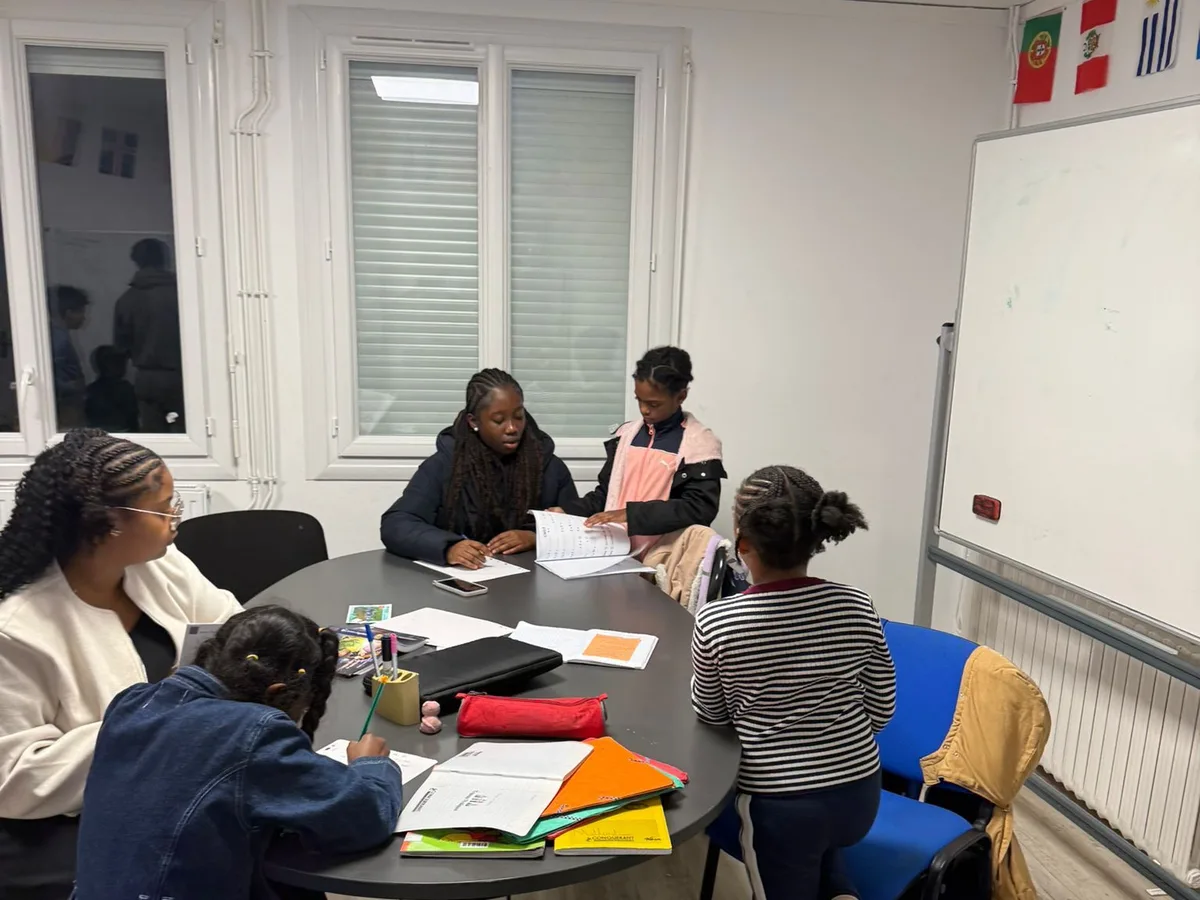 Autour d'une table de travail dans les locaux de l'AJN, deux bénévoles accompagnent quatre enfants pendant l'aide aux devoirs, cahiers et trousses ouverts.